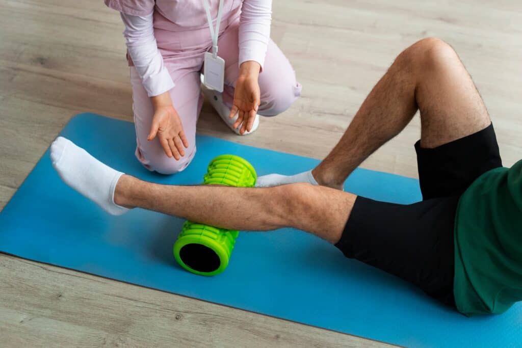 Una persona realiza terapia con un rodillo de espuma verde en la pierna en una colchoneta azul, mientras un fisioterapeuta la asesora y apoya en un entorno de gimnasio o clínica, promoviendo recuperación y bienestar físico.